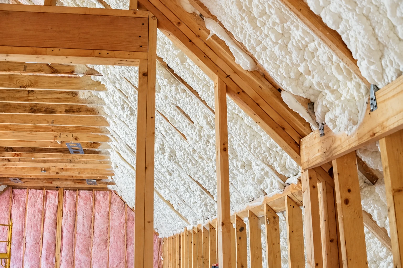 Exposed roof framing and rafters coated with white spray foam insulation, above pink batts in the lower wall.