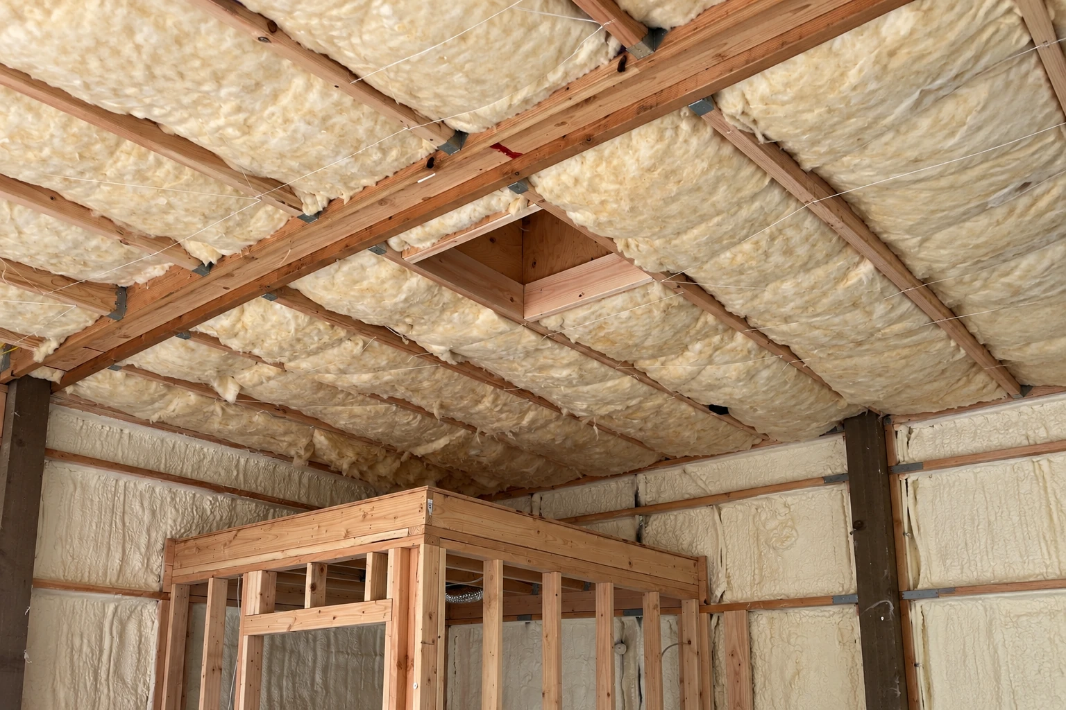 An unfinished attic has pale insulation packed between roof rafters and across the walls.
