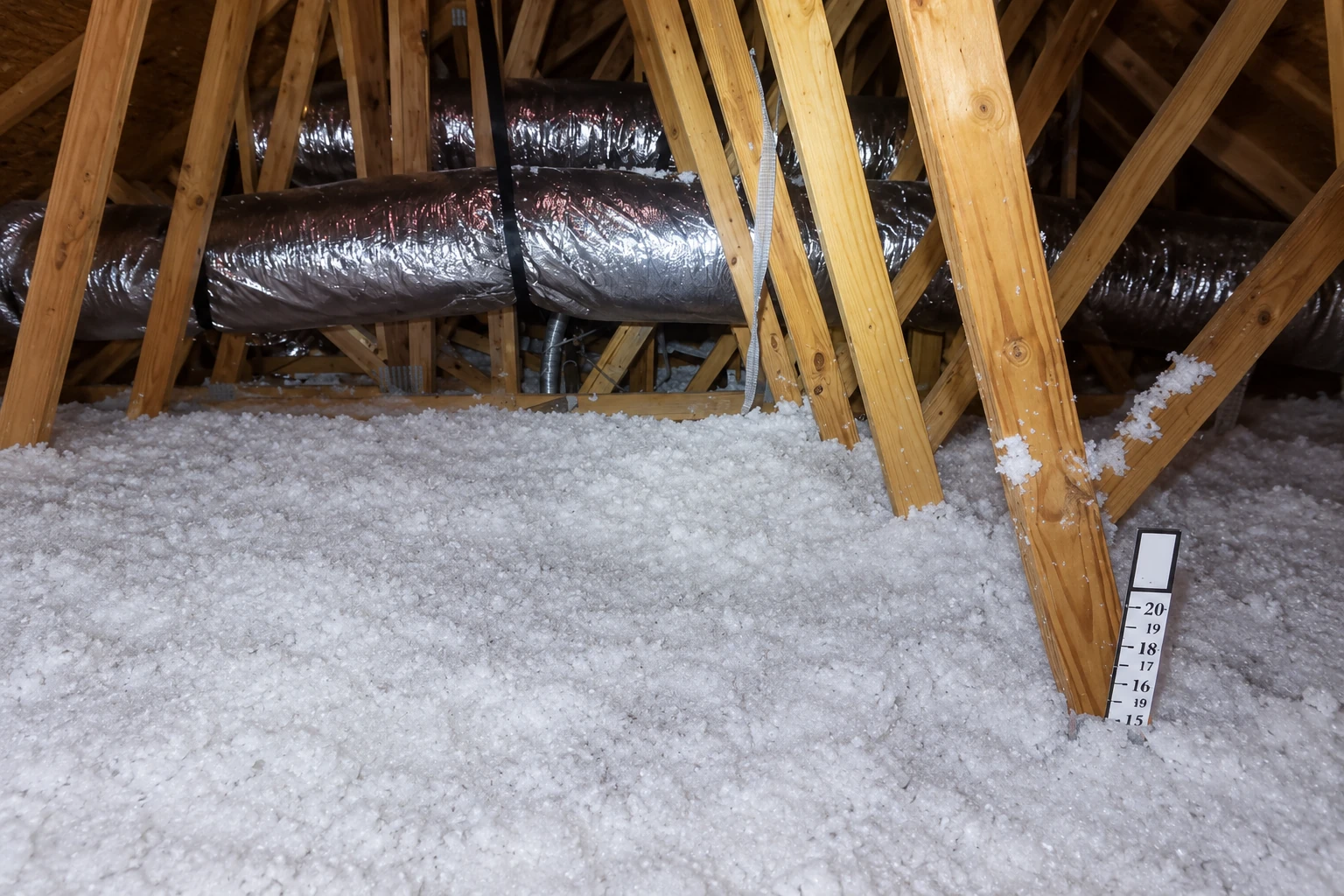 Loose white blown-in insulation covers an attic floor beneath wooden trusses and a metal duct.