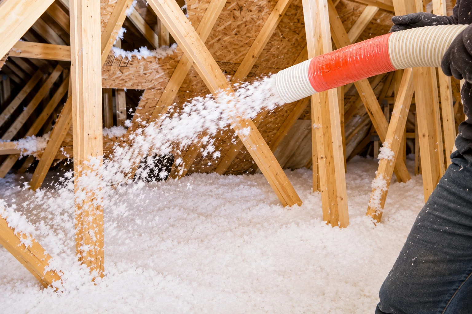 A hose blows white loose-fill insulation across the floor between exposed wooden attic trusses.
